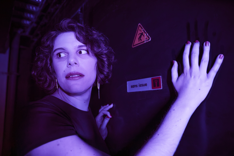 Portrait de Julia Popek. Elle pose les mains sur une armoire électrique et regarde derrière elle. Elle est dans les ténèbres, seulement illuminée par une lumière bleue.